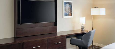 Habitación de hotel con un televisor de pantalla plana grande montado en un mueble de madera oscura, un escritorio con una silla, una lámpara y un cuadro abstracto enmarcado en la pared.