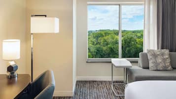 A hotel room with a bed, a desk with a lamp, a chair, a sofa with a pillow, a window with a view of trees, and a carpeted floor.