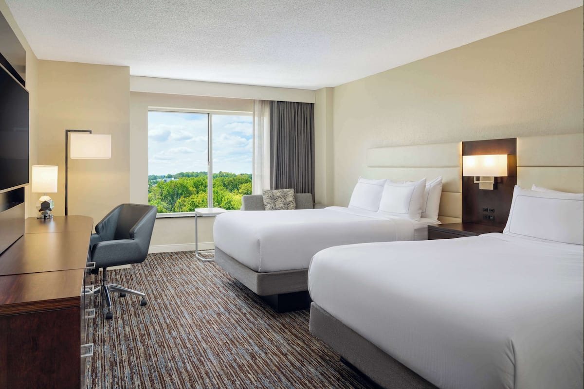 A hotel room with two beds, a desk, a chair, and a large window offering a view of greenery.