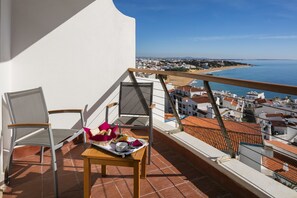 Un balcón con dos sillas, una mesa con comida y vistas al mar y edificios.