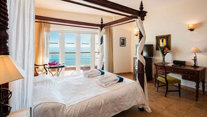 Una cama con dosel con sábanas blancas, un escritorio con lámpara y vistas al mar a través de amplias ventanas.
