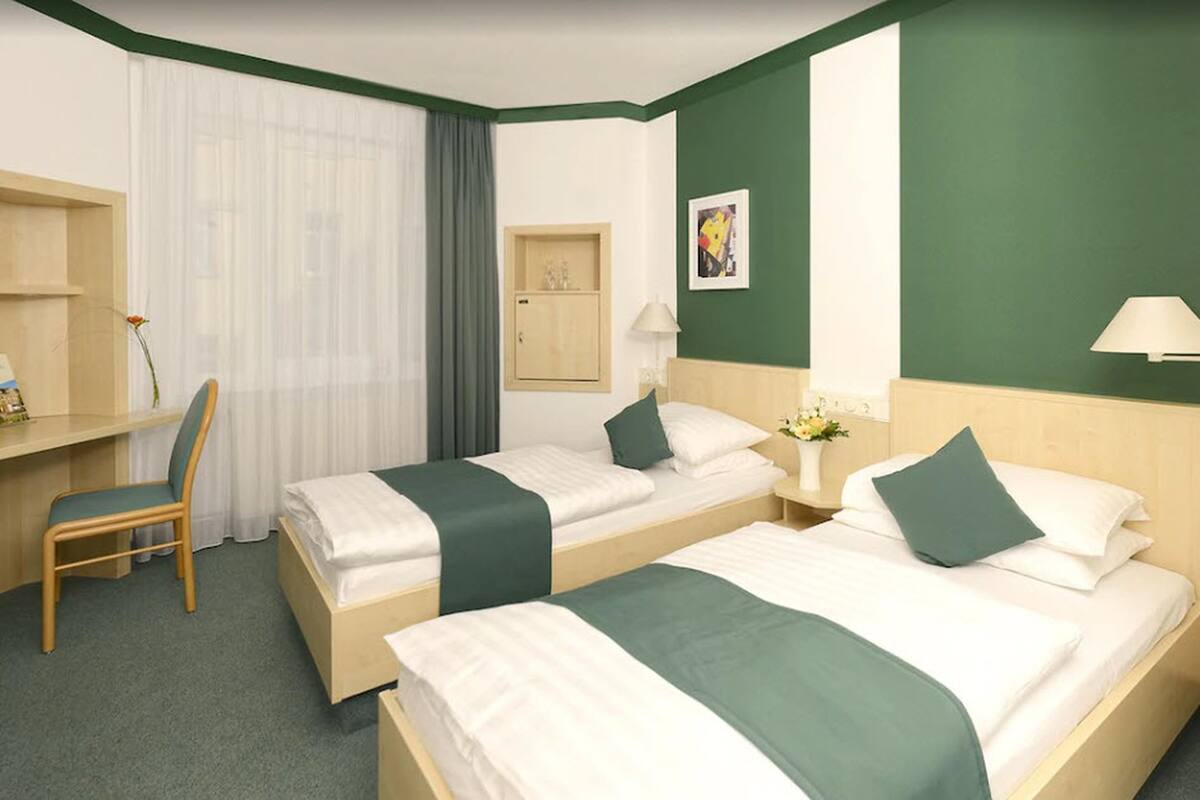 A hotel room with two beds, a desk, a chair, a small cabinet, and a window with curtains.