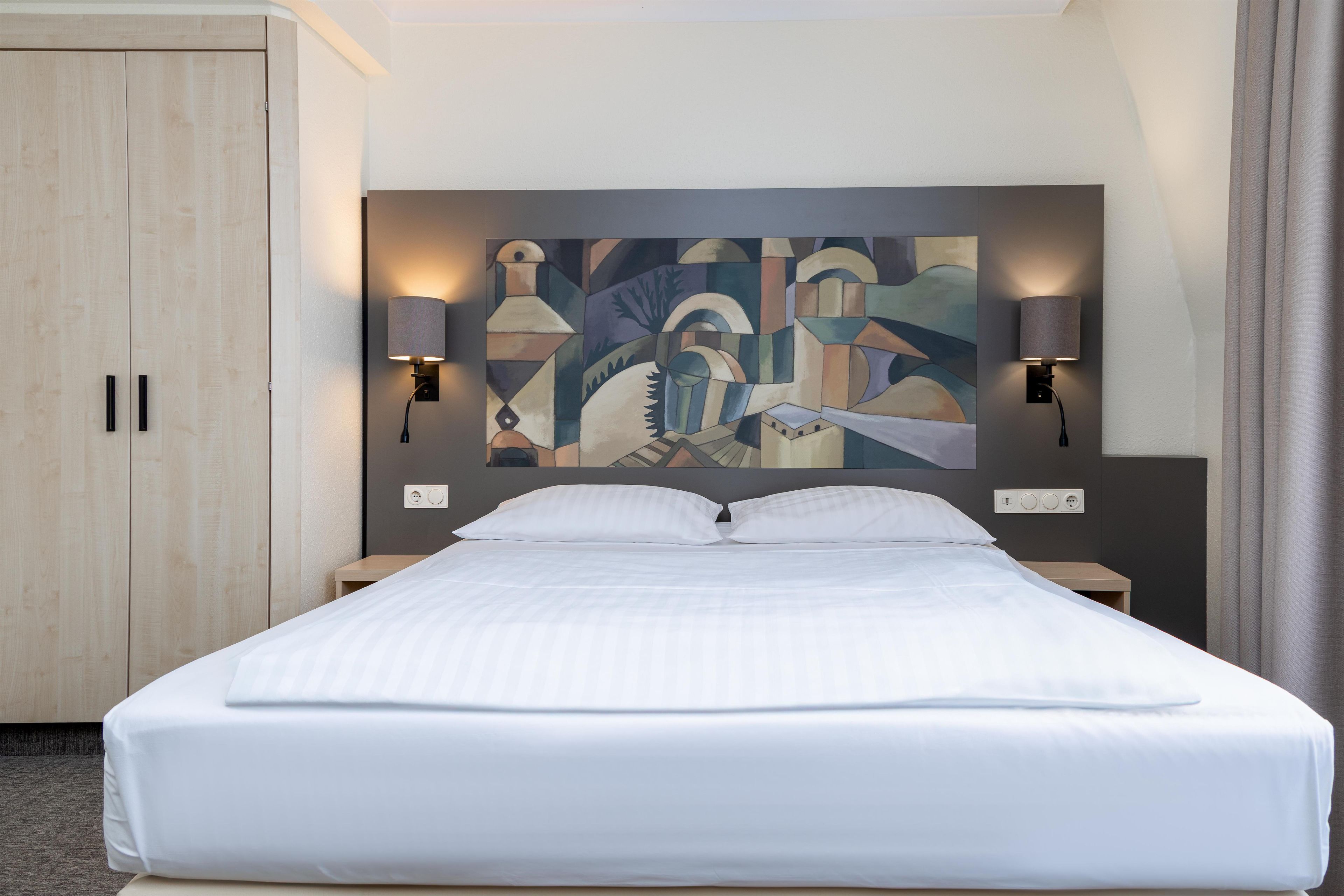 A hotel room with a bed, a headboard with a painting, two wall-mounted lamps, and a wardrobe.