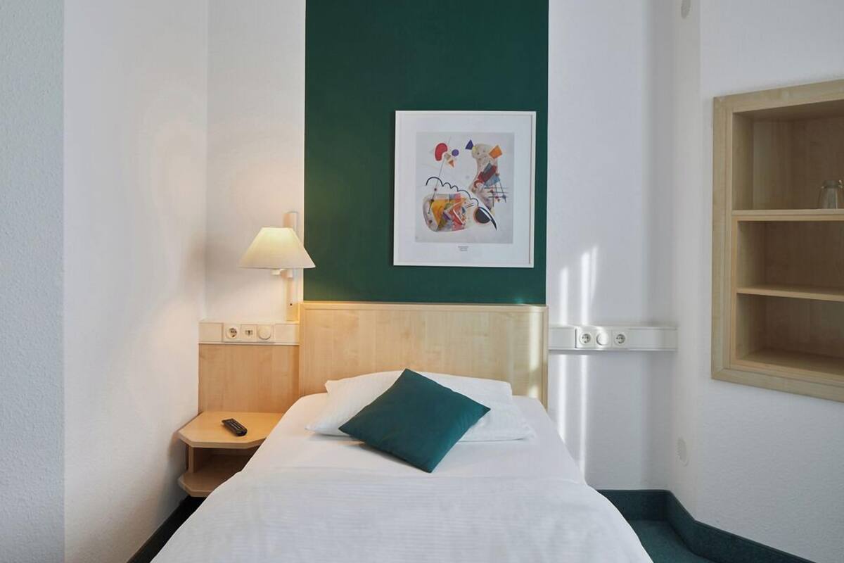 A hotel room with a bed, a nightstand, a lamp, a wall-mounted picture, and a wooden shelf.