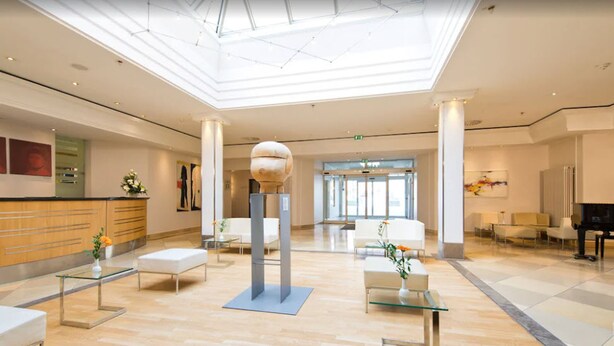 A spacious lobby with a high ceiling, a reception desk, seating areas, and a large decorative sculpture. - Hotel Kaiserin Augusta (Weimar)