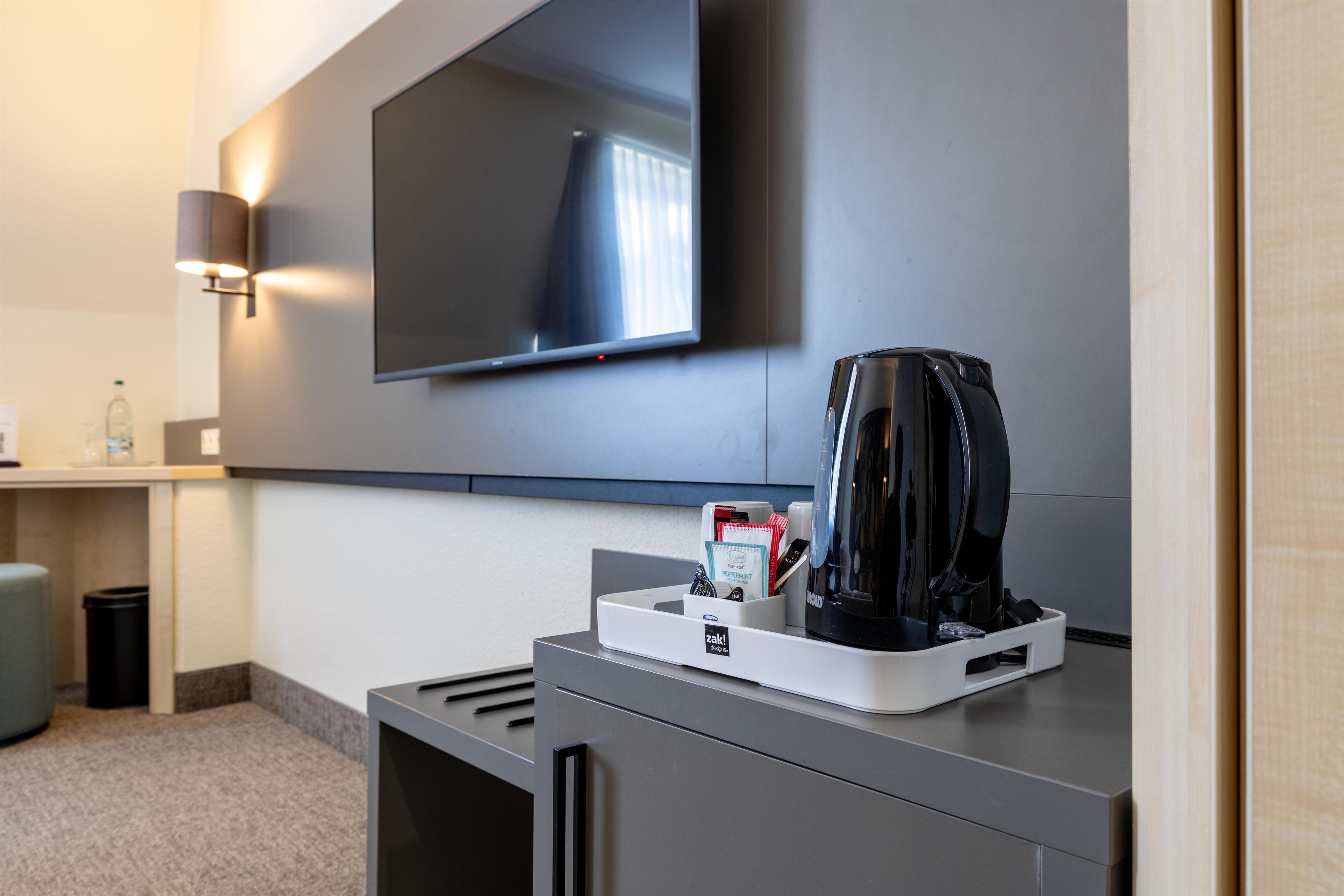 A modern hotel room with a flat-screen TV mounted on the wall, a coffee maker on a counter, and a trash bin.