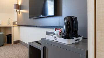 A modern hotel room with a flat-screen TV mounted on the wall, a coffee maker on a counter, and a trash bin.