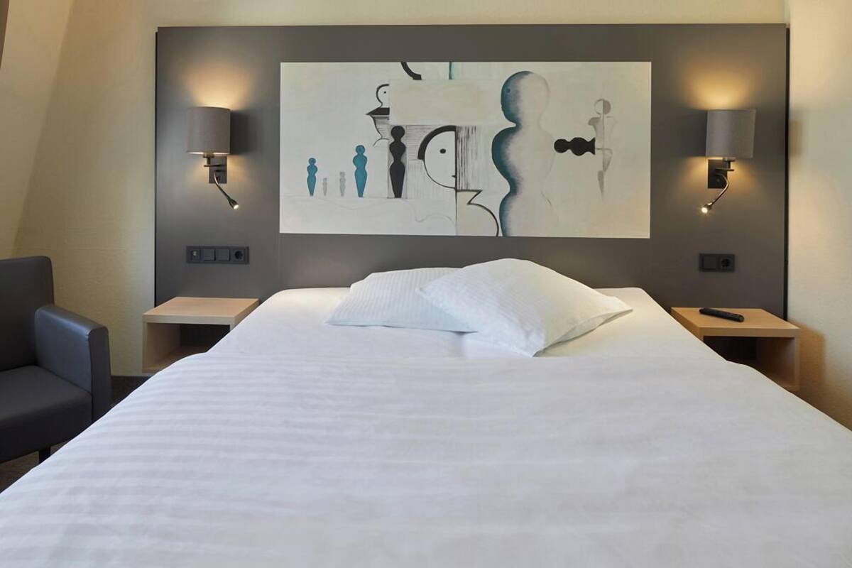 A hotel room with a large bed, two bedside tables, a chair, and a wall-mounted abstract artwork.