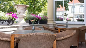 Un'area salotto esterna con mobili in vimini, un vaso con fiori e un'ampia finestra vista sugli edifici.