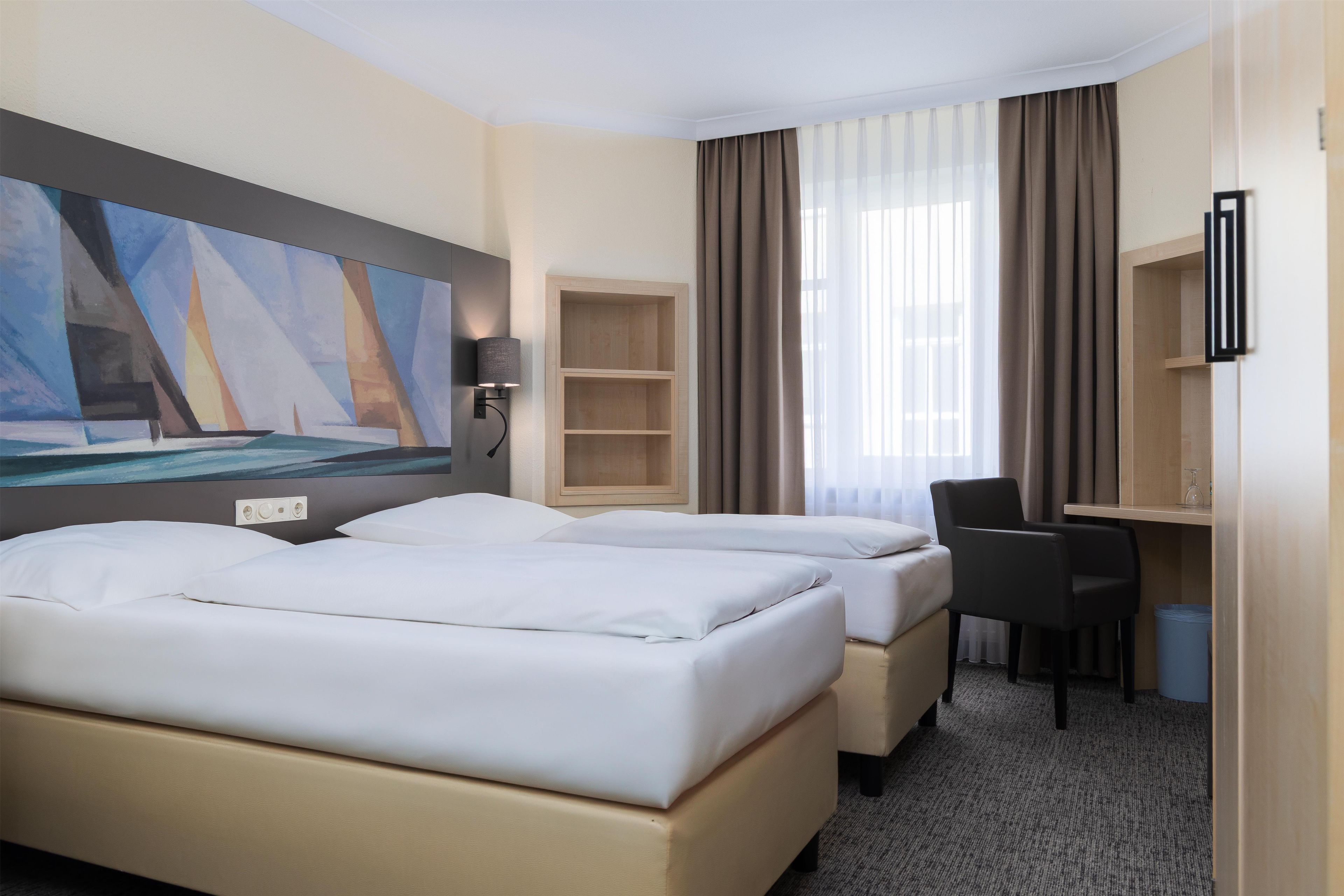 A hotel room with two beds, a desk, a chair, a painting, and a window with curtains.