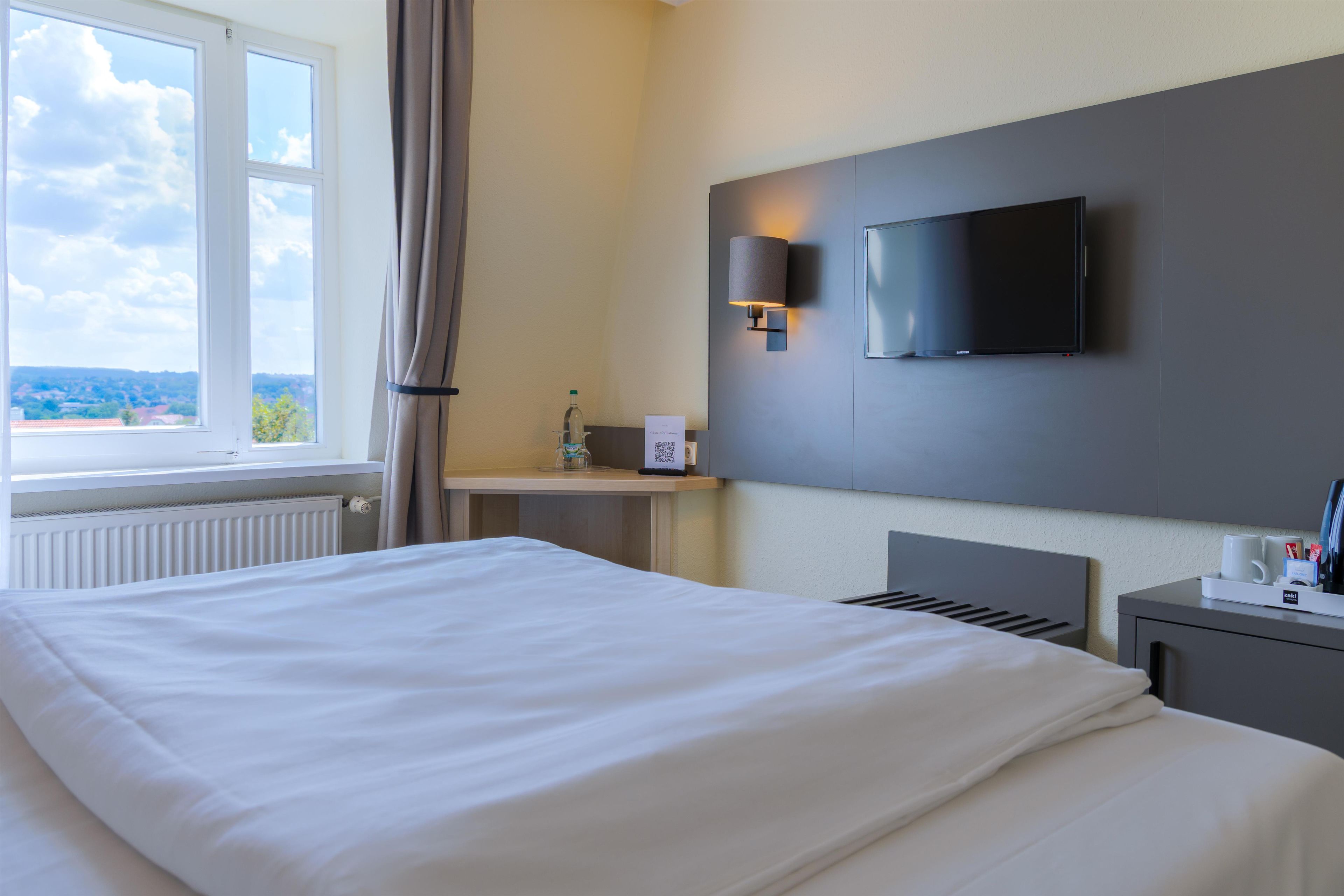 A hotel room with a large bed, a flat-screen TV mounted on the wall, a bedside table with a lamp, and a window with curtains.