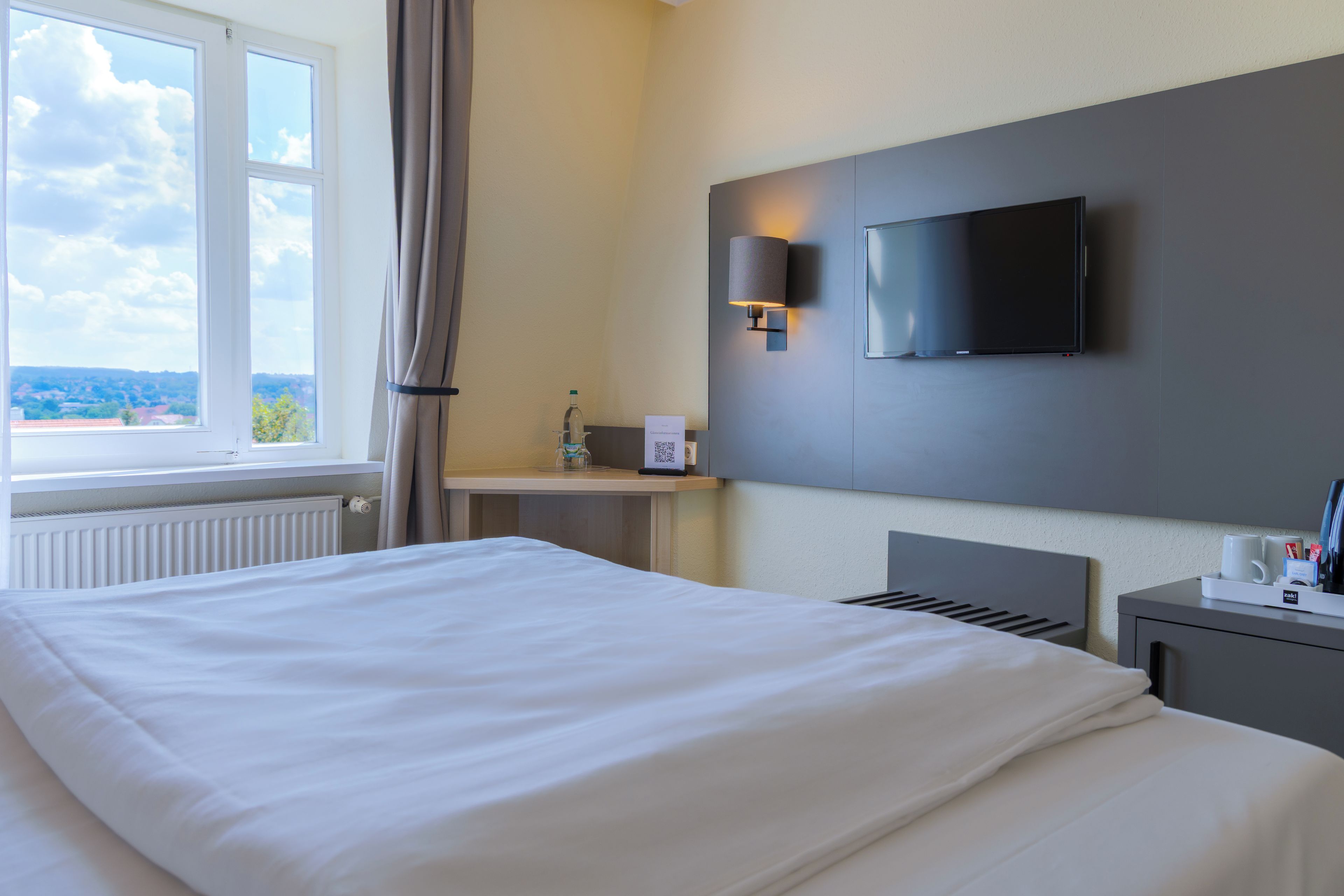 A hotel room with a bed, a TV mounted on the wall, a bedside table with a lamp, and a window with curtains.
