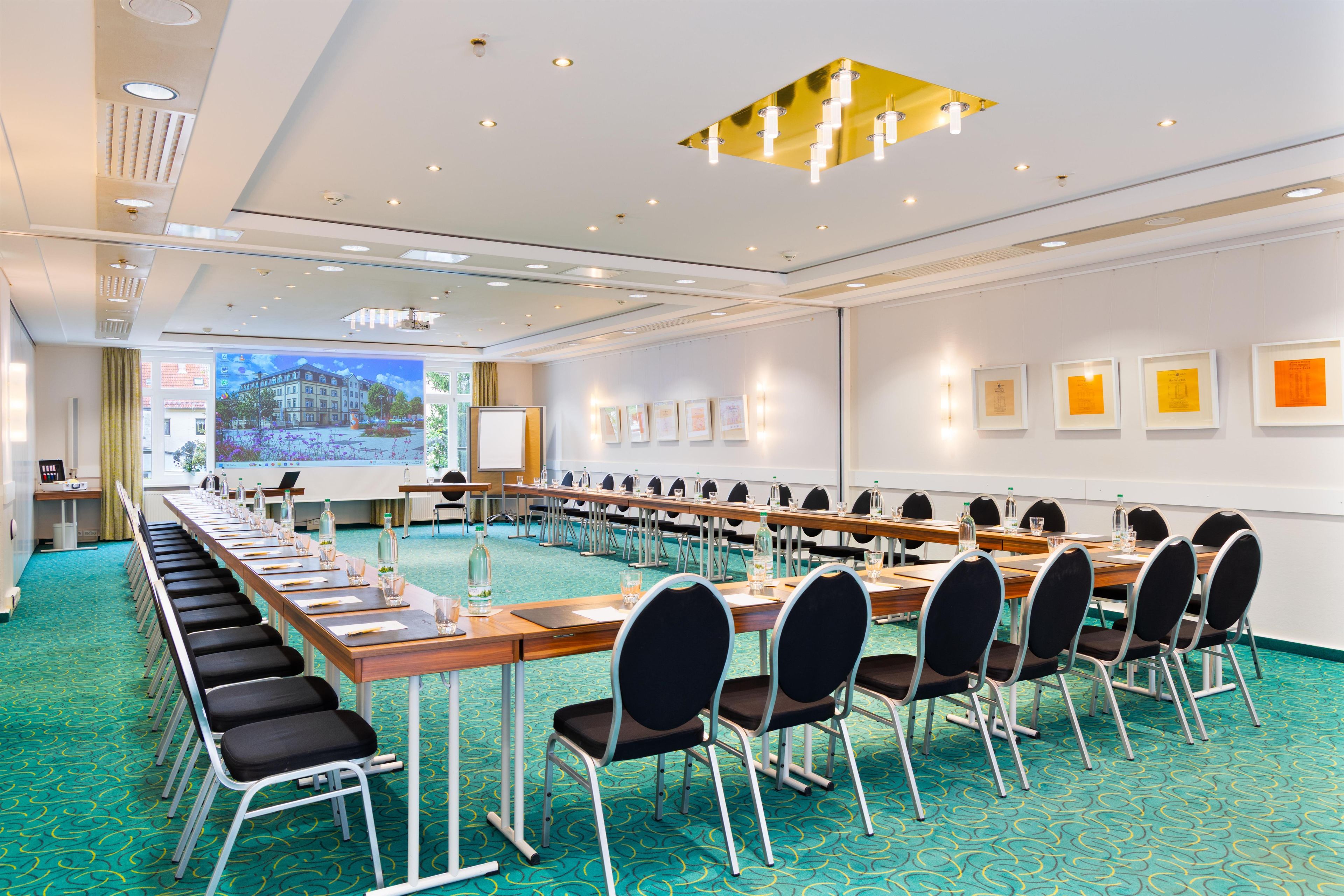 Ein Konferenzraum mit einem langen Tisch, Stühlen, einer Projektionsleinwand und gerahmten Bildern an der Wand.