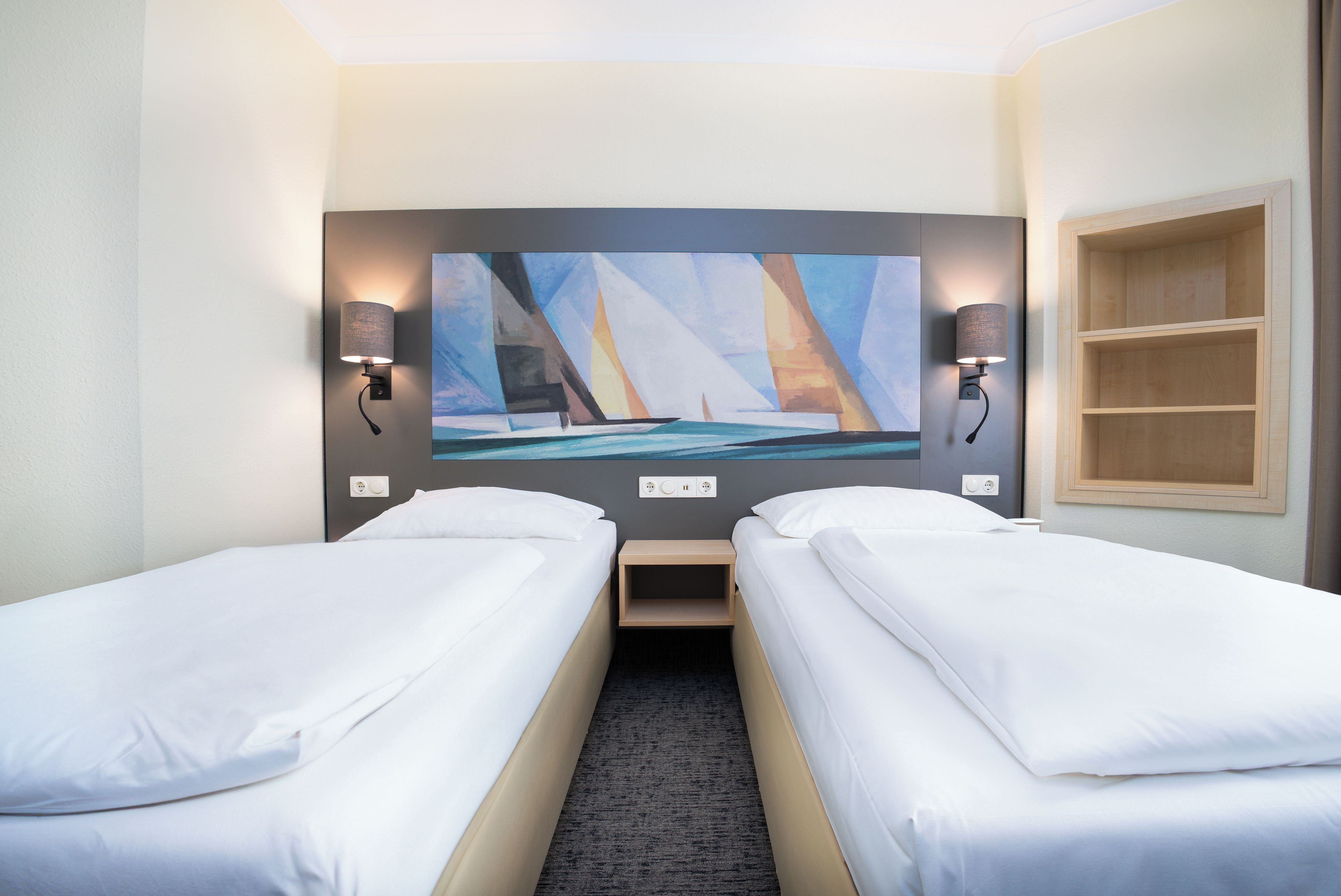 Een hotelkamer met twee eenpersoonsbedden, een schilderij van zeilboten aan de muur en een houten plankunit.