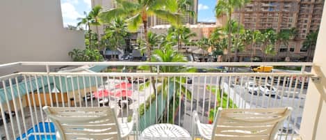 A balcony with two white chairs and a small round table, overlooking a street with palm trees and tall buildings.