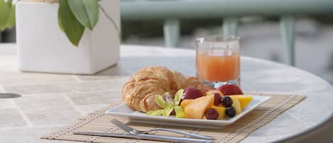 A table with a croissant, fruit, and a glass of orange juice, a potted orchid, and a view of the city and ocean.