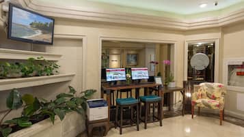 A hotel lobby with a reception desk, two monitors, a printer, and a comfortable seating area with a chair and stools.