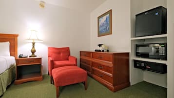 Una habitación de hotel con una cama, un sillón rojo, una mesita de noche de madera con una lámpara, un aparador, un microondas y un cuadro en la pared.