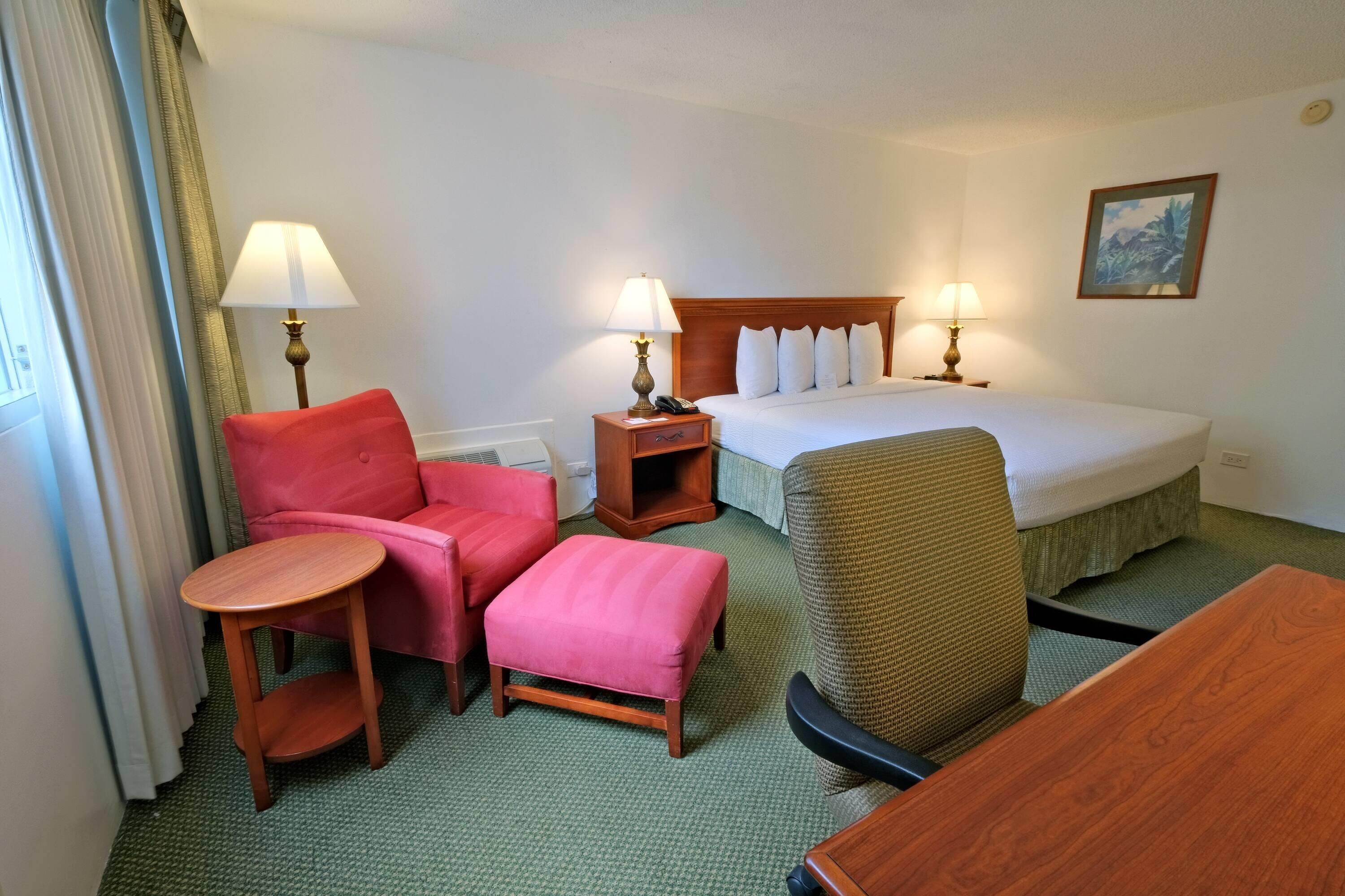 A hotel room with a bed, two bedside tables with lamps, a small round table, a red armchair, a brown armchair, and a green carpet.