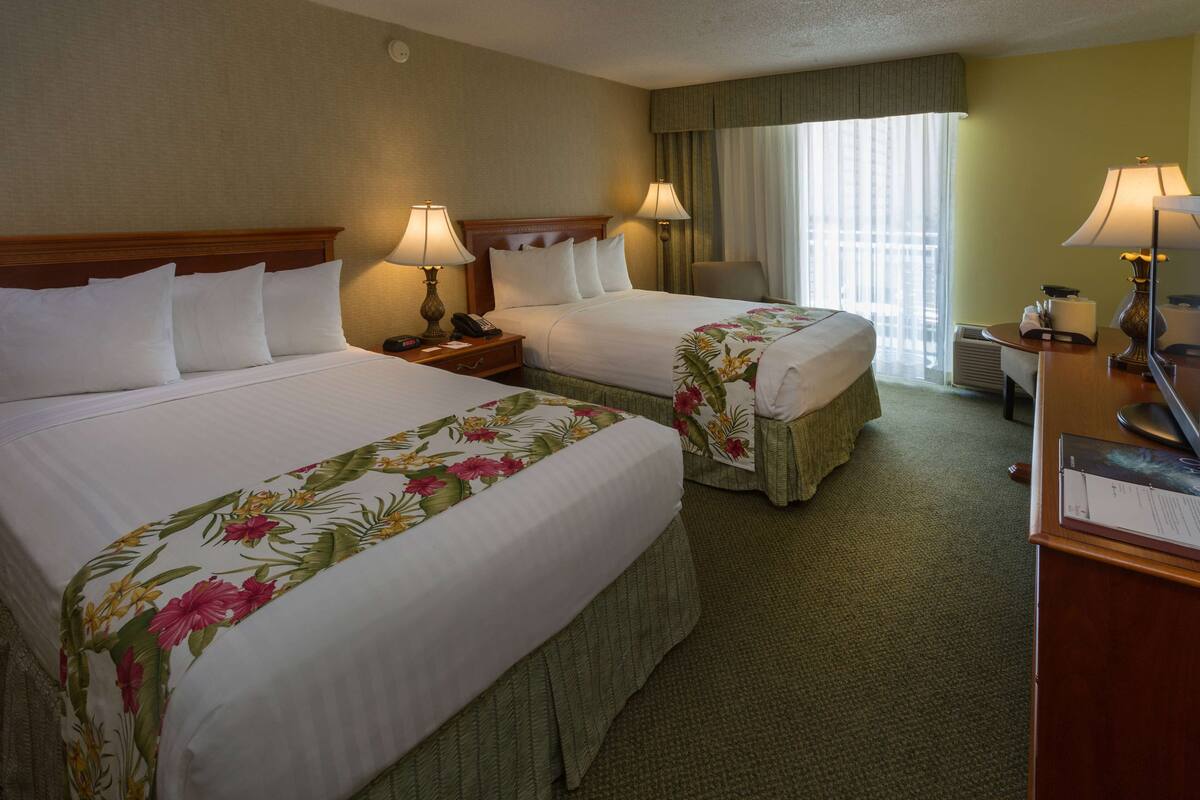 Een hotelkamer met twee bedden, elk voorzien van een bloemenprint sprei, nachtlampjes, een bureau met een computer en een raam met gordijnen.