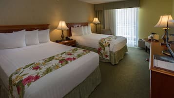 Quarto de hotel com duas camas, cada uma com colchas florais, luminárias laterais, uma escrivaninha com computador e uma janela com cortinas.