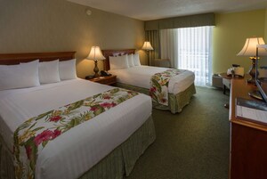 A hotel room with two beds, each with floral bedspreads, bedside lamps, a desk with a computer, and a window with curtains.