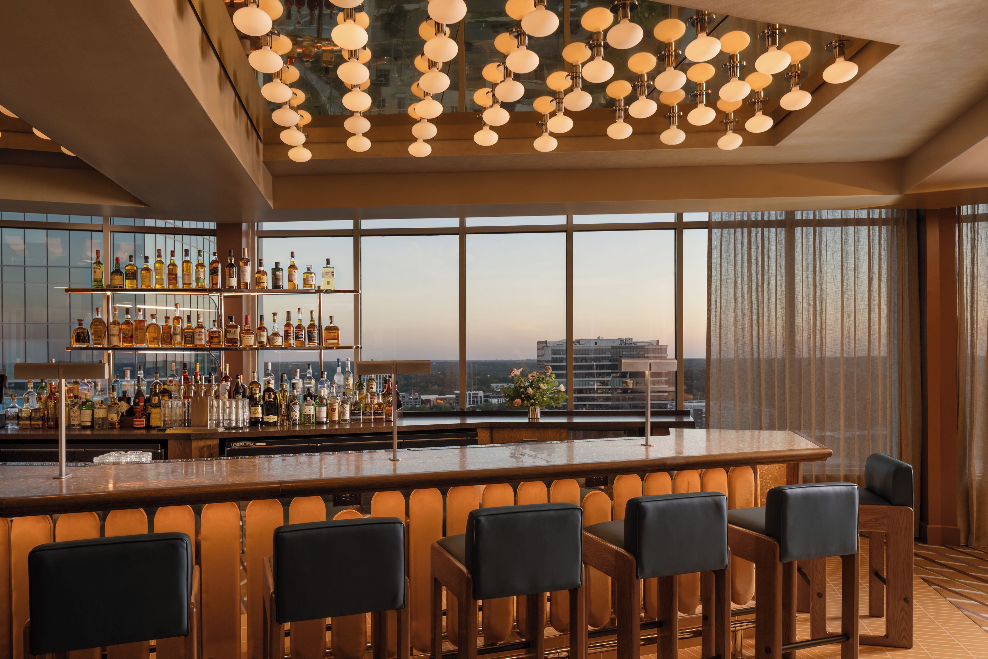 A bar area with a long counter, multiple bottles, and a view of buildings through large windows.
