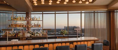 A bar area with a long counter, multiple bottles, and a view of buildings through large windows.
