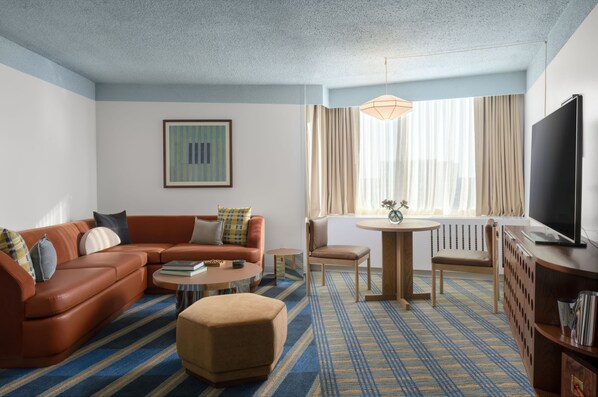 Une chambre d’hôtel comprenant un canapé, un pouf, une table basse, une télévision et un coin repas avec une table et des chaises.