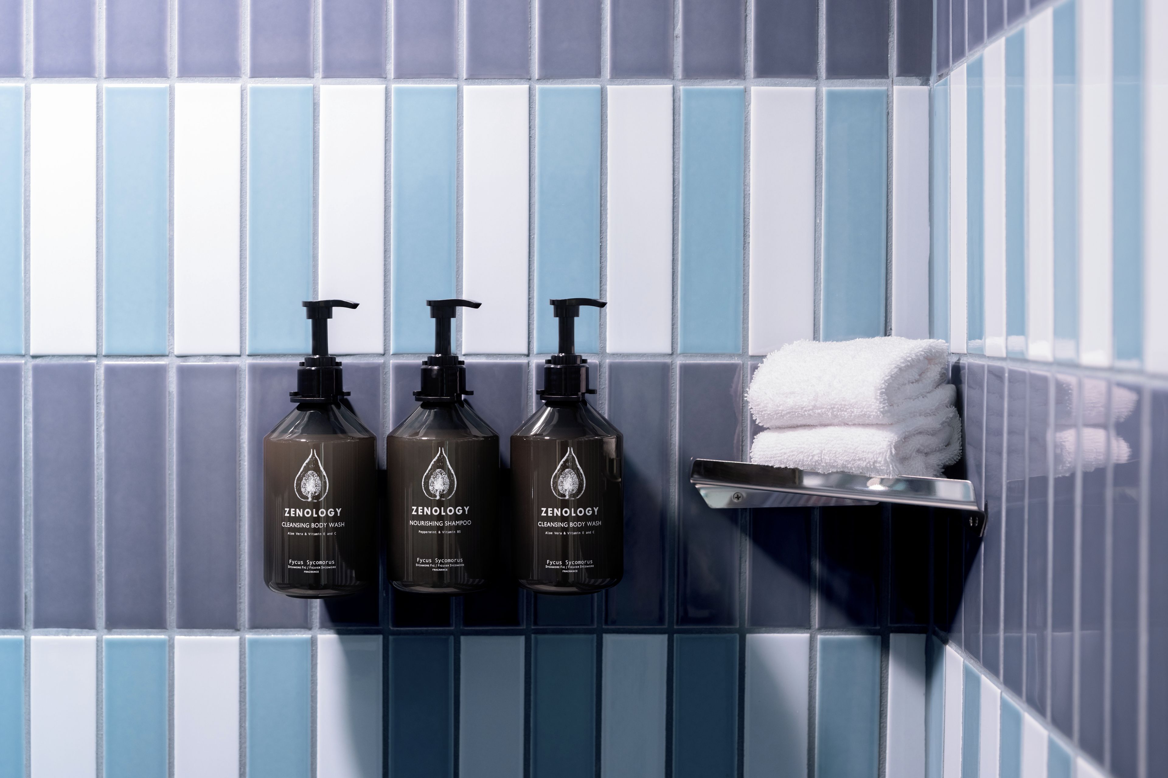 A bathroom with three bottles of Zenology Nourishing Shampoo and Cleansing Body Wash, a white towel on a rack, and a blue and white tiled wall.