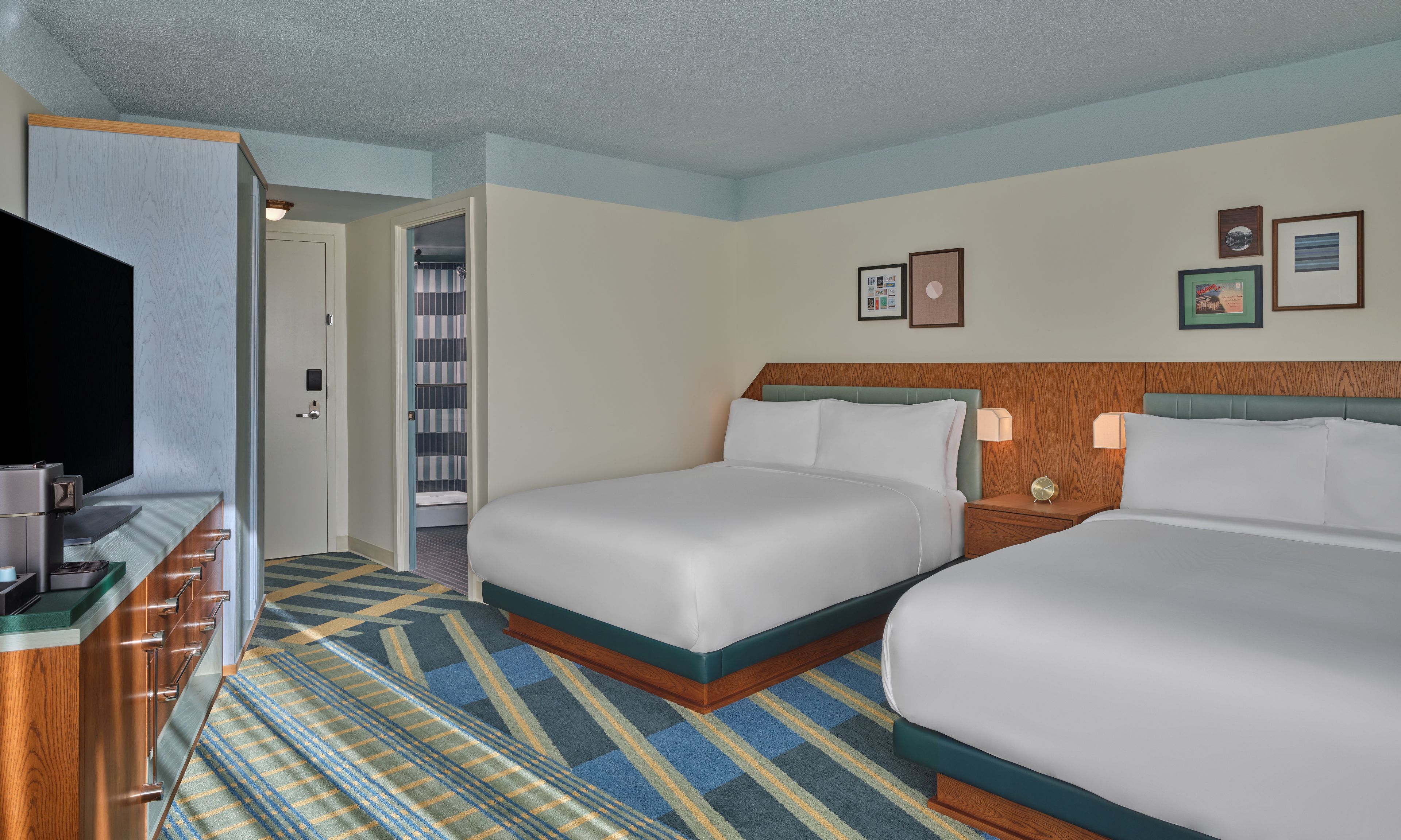 A hotel room with two beds, a television, a desk with a computer, and a bathroom visible through an open door.