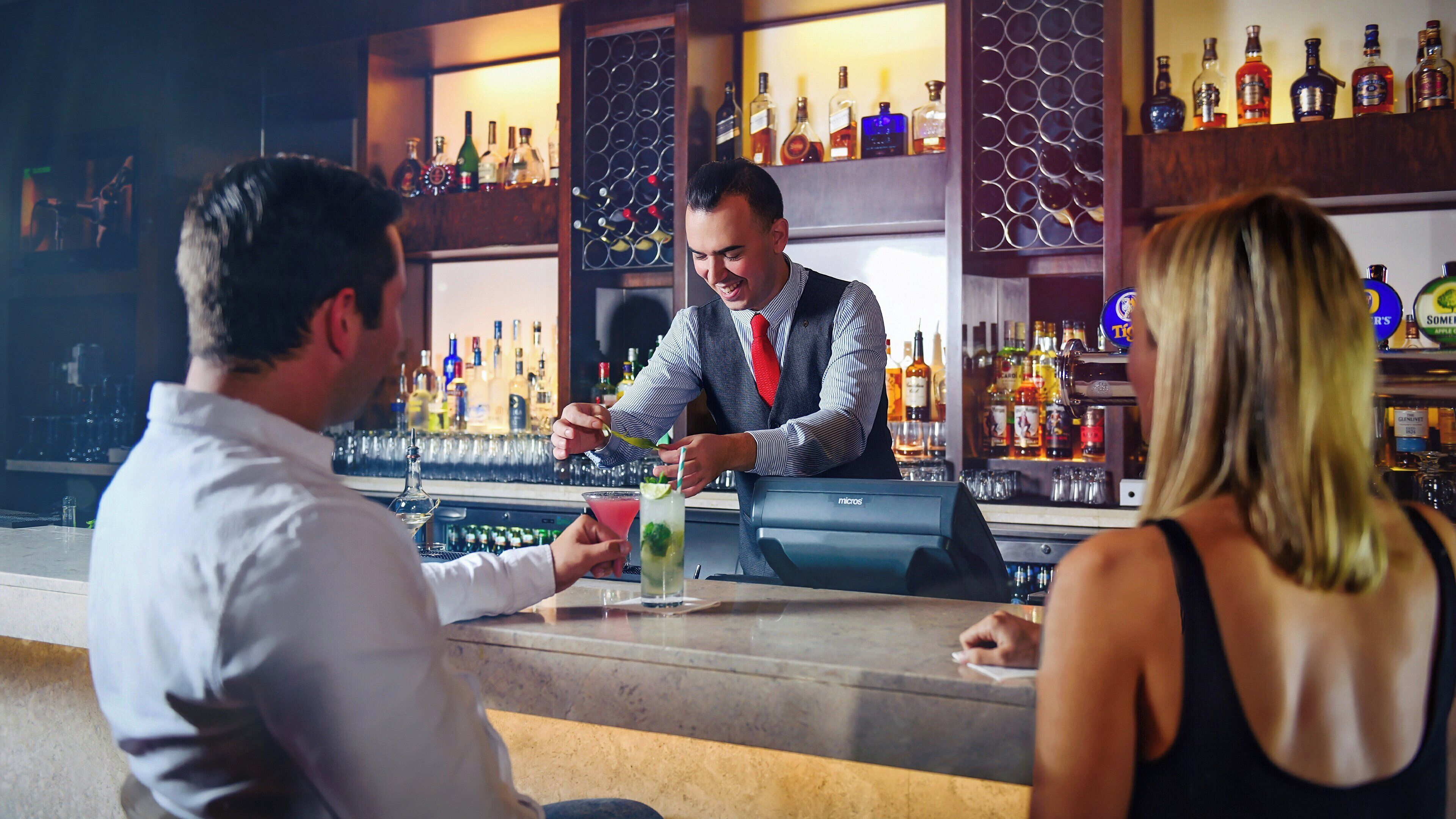 Un bar con un cantinero preparando una bebida. Hay estanterías con diferentes botellas y vasos detrás de la barra.
