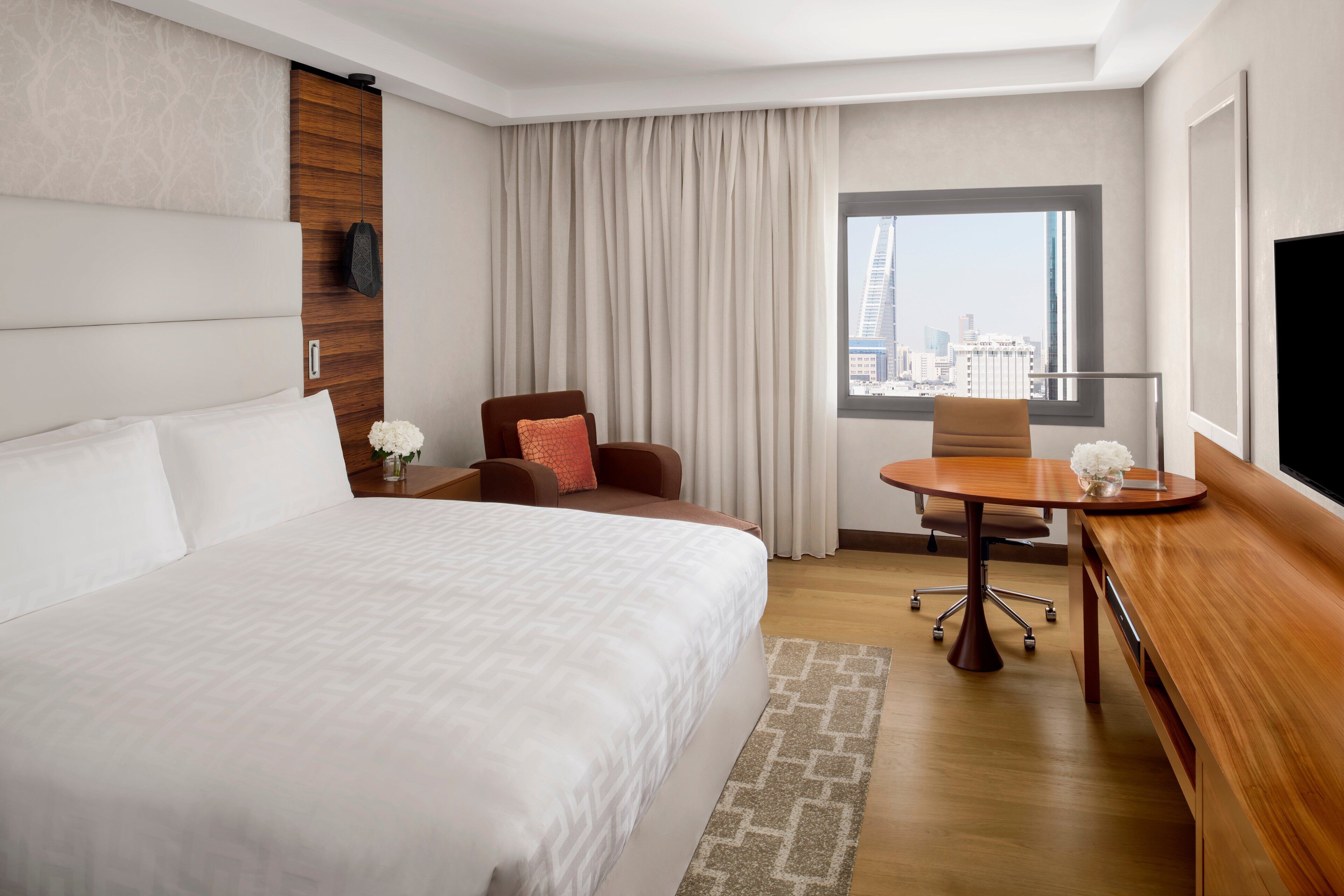 A hotel room with a large bed, a desk, a chair, a small table, and a view of the city.