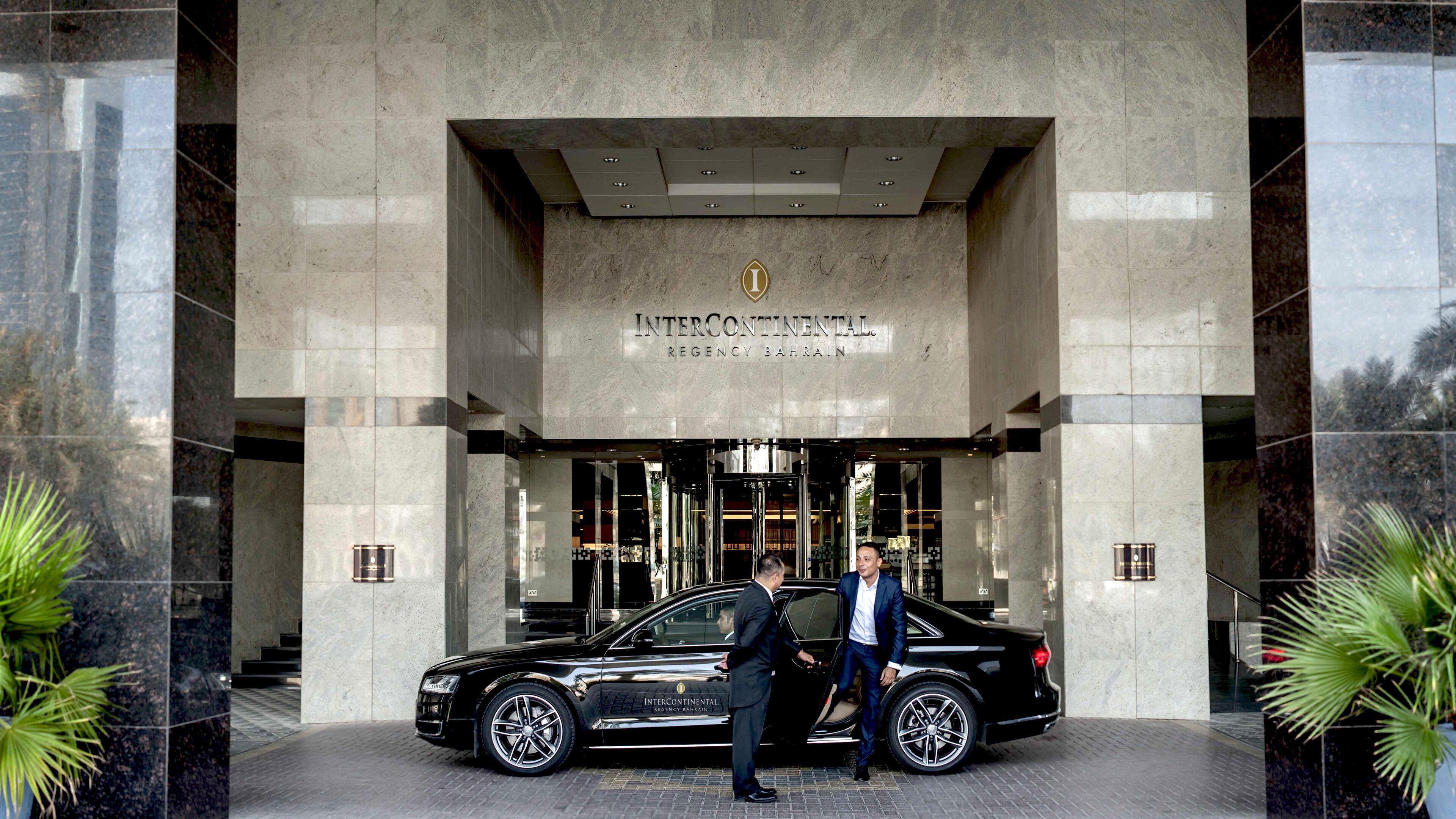 Un coche de lujo negro estacionado frente a un edificio de fachada de mármol, un letrero que dice 'InterContinental Regency Bahrain', y dos hombres de pie junto al coche.
