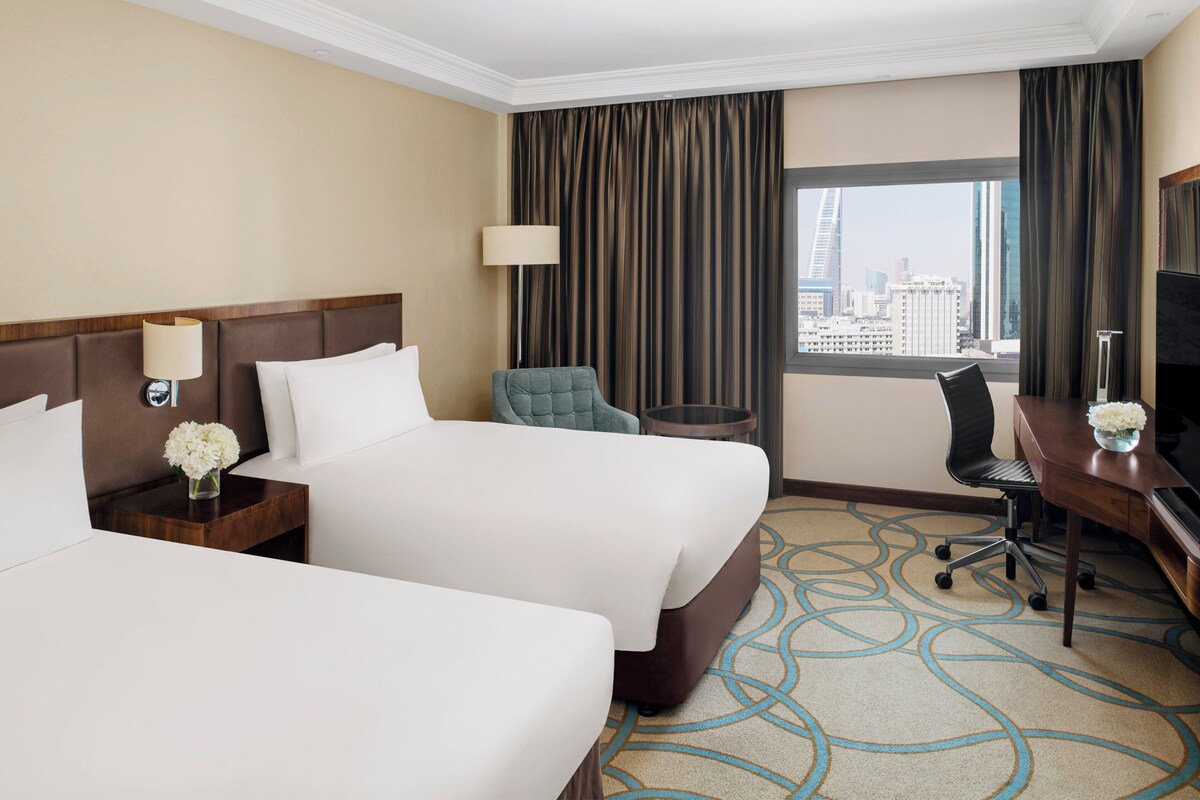 A hotel room with two beds, a desk, a chair, a TV, a lamp, and a view of the cityscape.