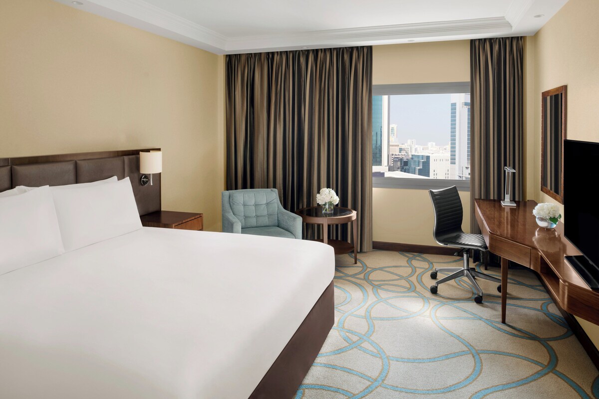 A hotel room with a large bed, a desk with a chair, a small table with a vase, and a TV.