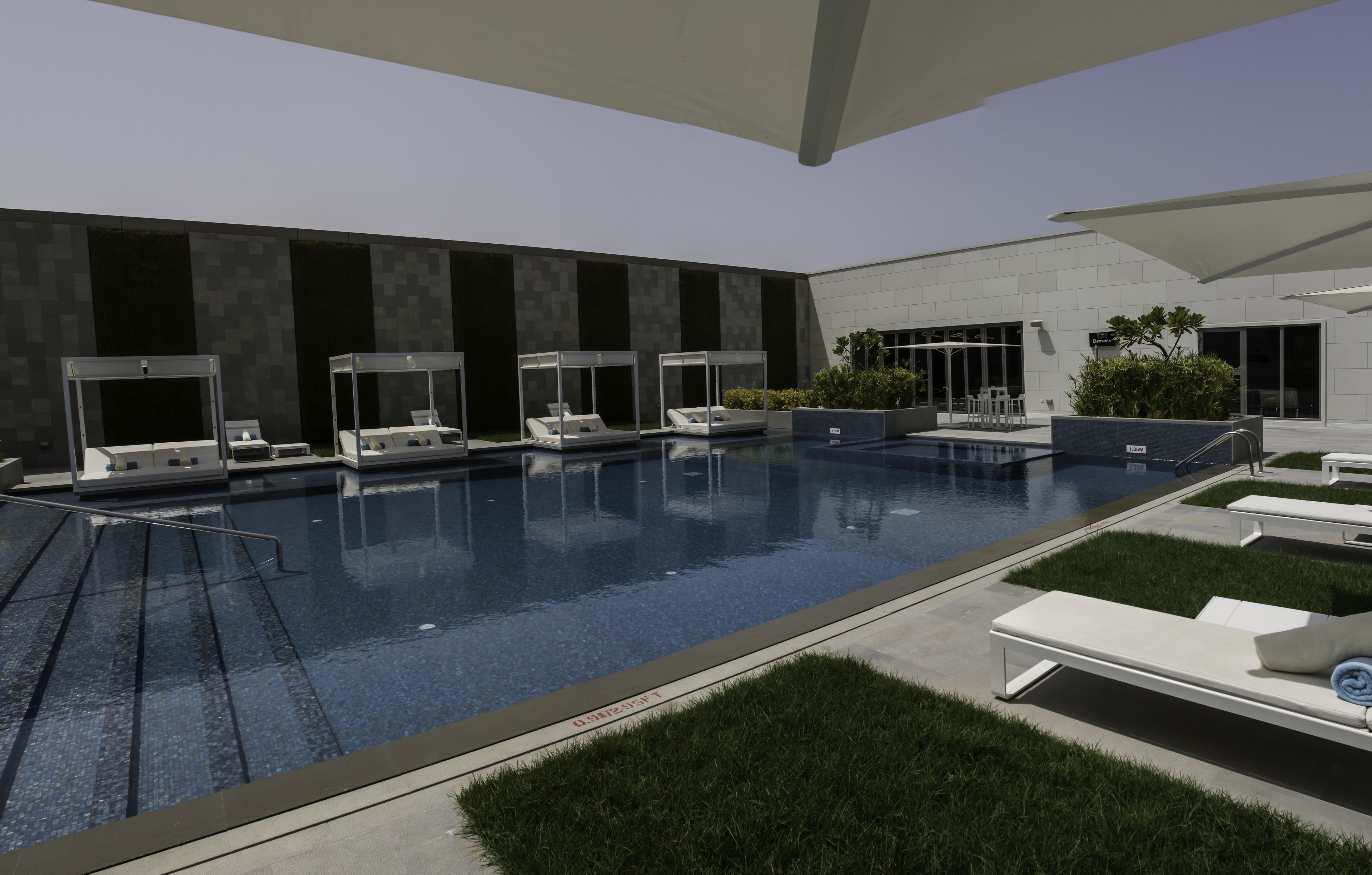 A modern indoor pool area with lounge chairs and sunbeds, surrounded by a landscaped garden and a building with large windows.