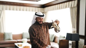 A man in traditional attire pouring coffee into a cup in a well-lit room with a sofa, coffee table, and lamp.