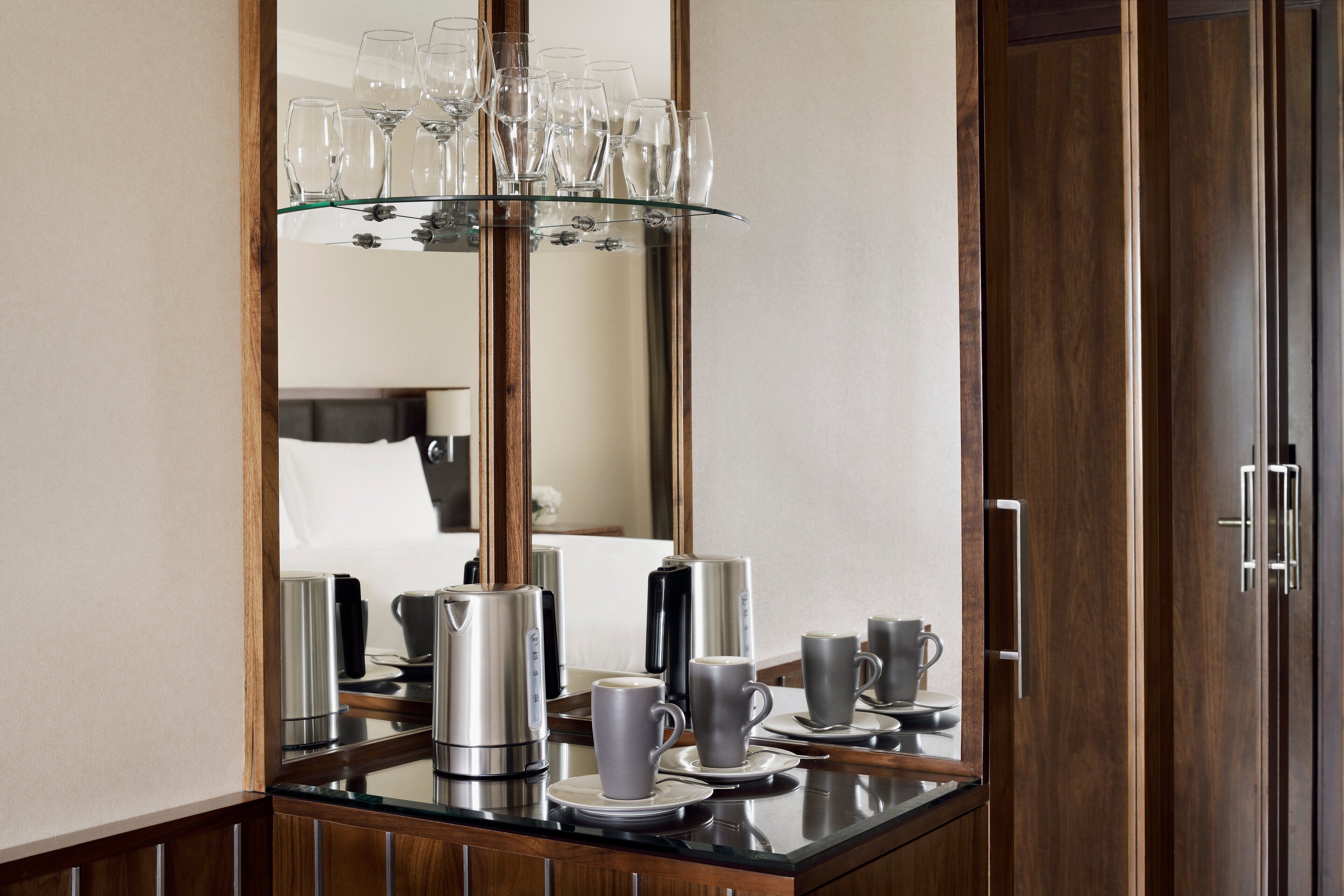 A hotel room with a mirrored wardrobe, a glass coffee table with a tea set, and a bed with a white headboard.