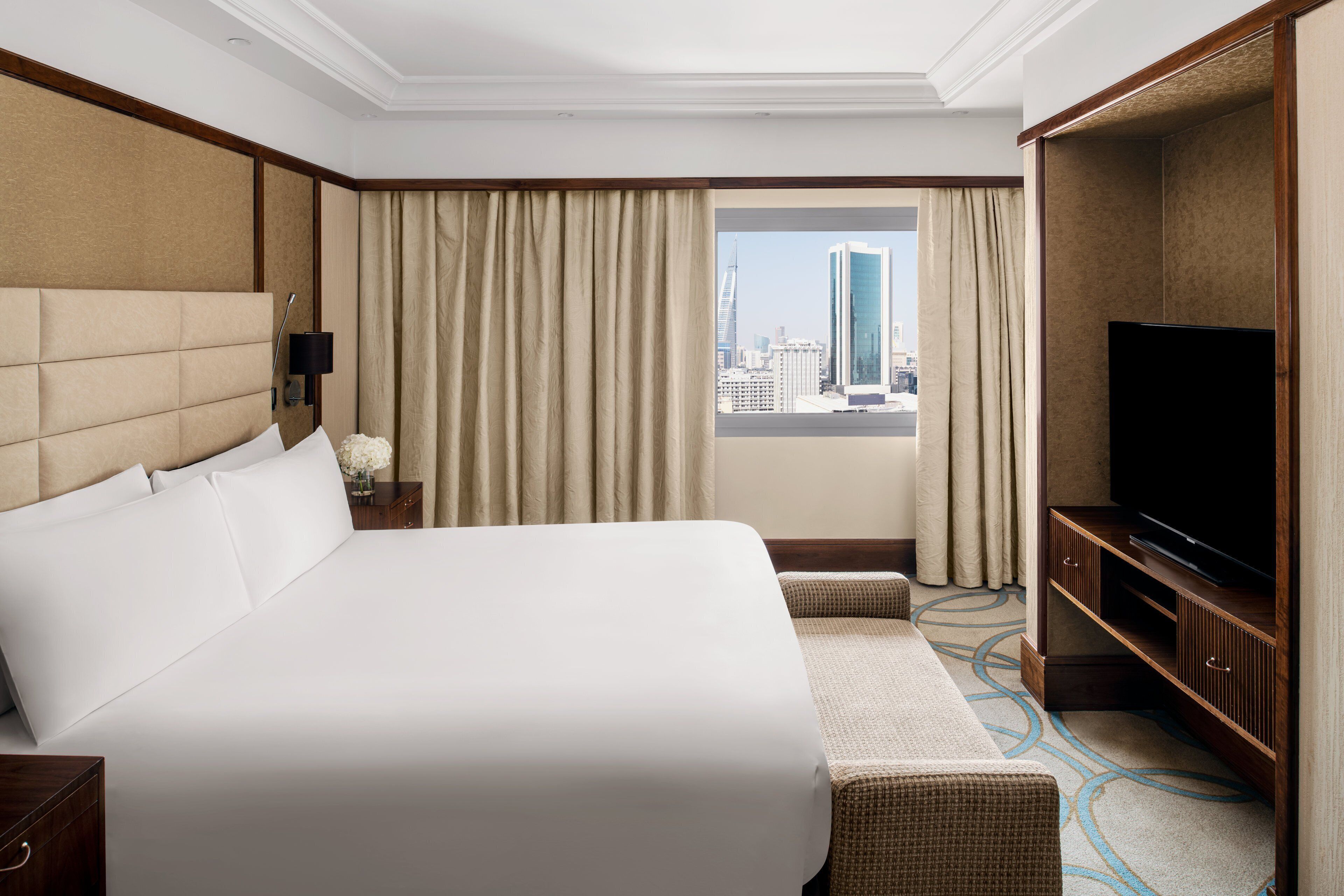 A hotel room with a large bed, a television, a nightstand, and a view of a cityscape through the window.