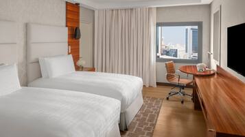 A hotel room with two beds, a desk, a chair, a TV, and a view of the city.