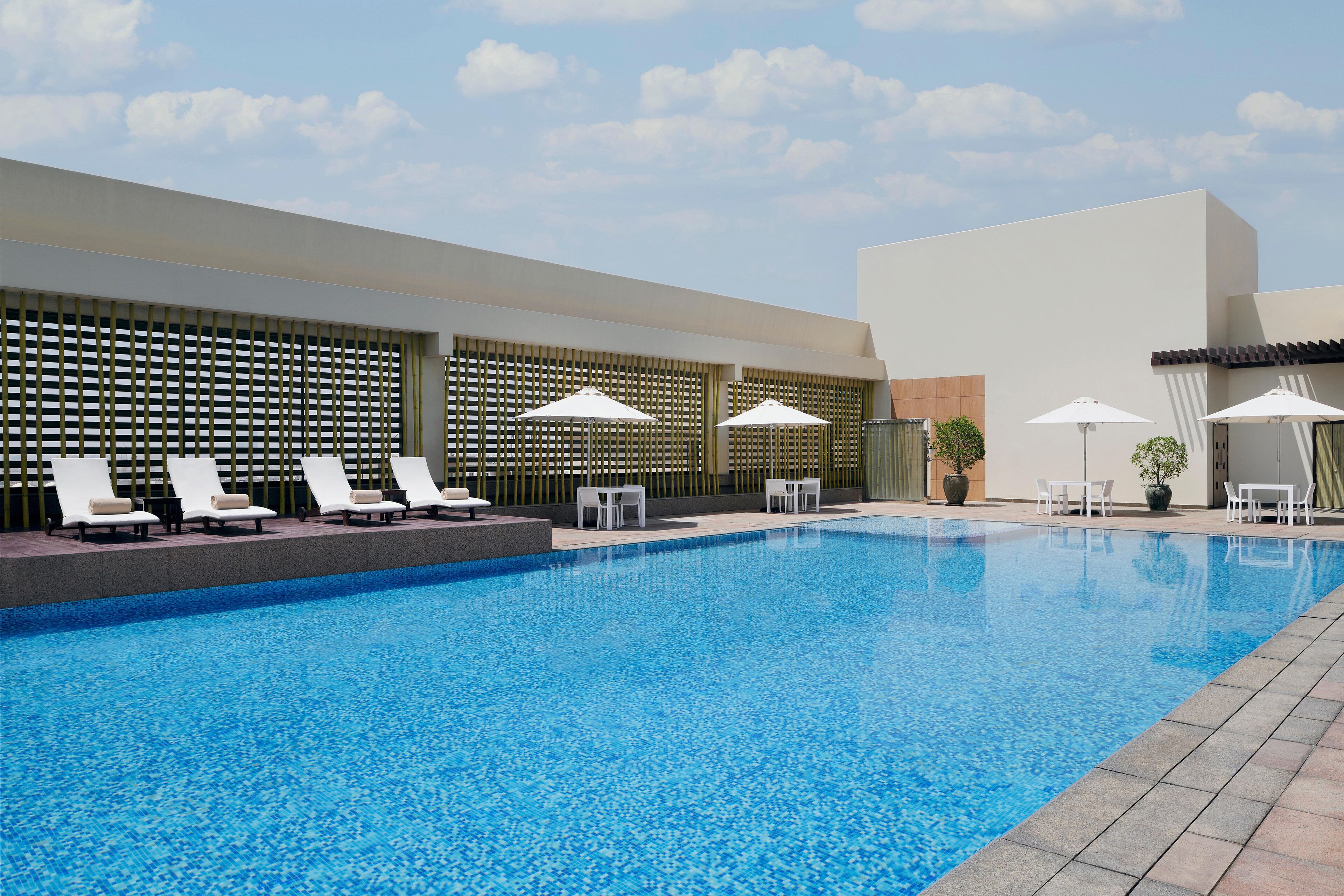 Una piscina con sillones de playa y sombrillas, rodeada por un edificio moderno con suelo de baldosas.