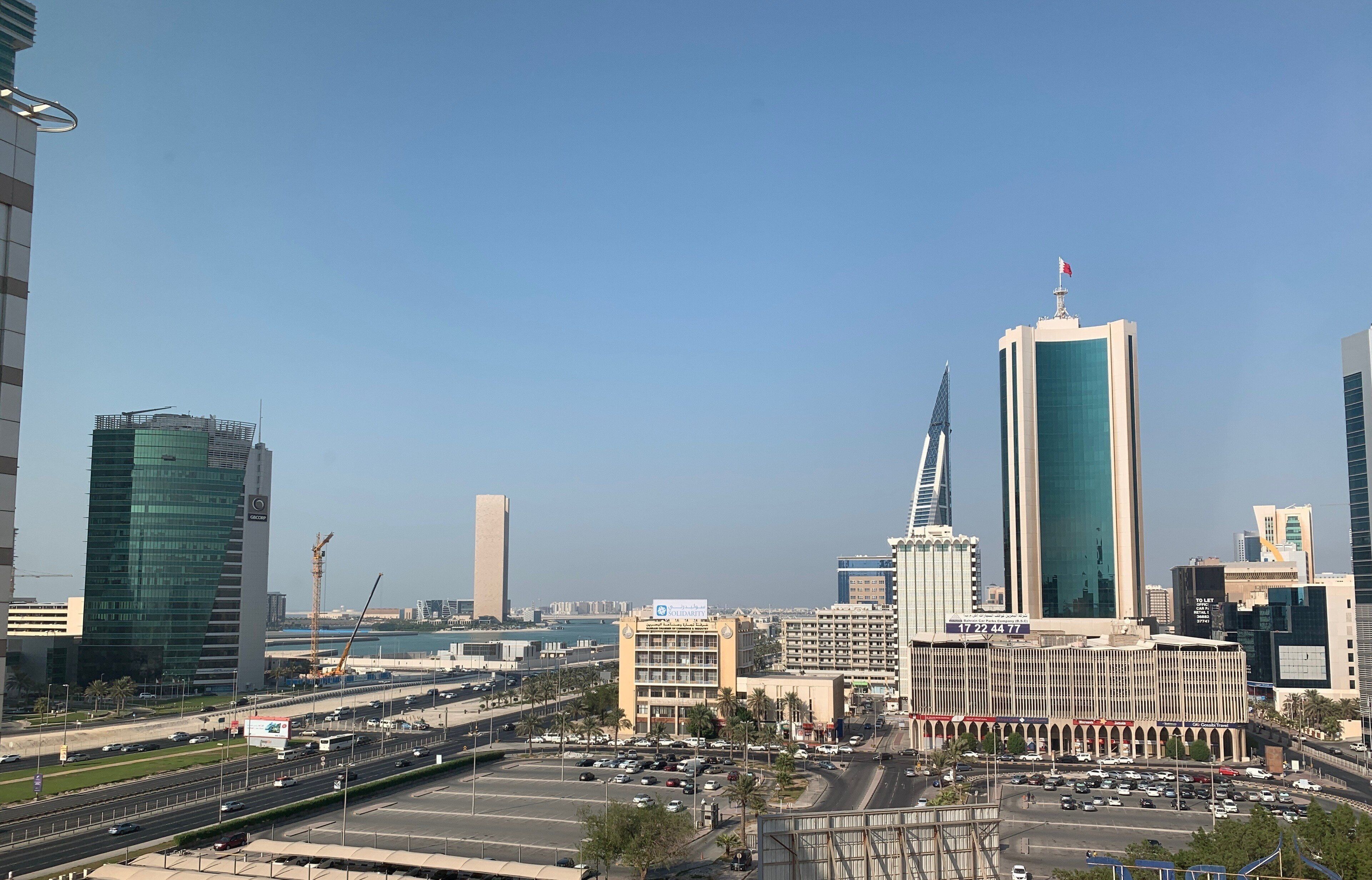 A cityscape with modern high-rise buildings, a prominent tower with a flag, and a busy intersection with vehicles and a parking lot.