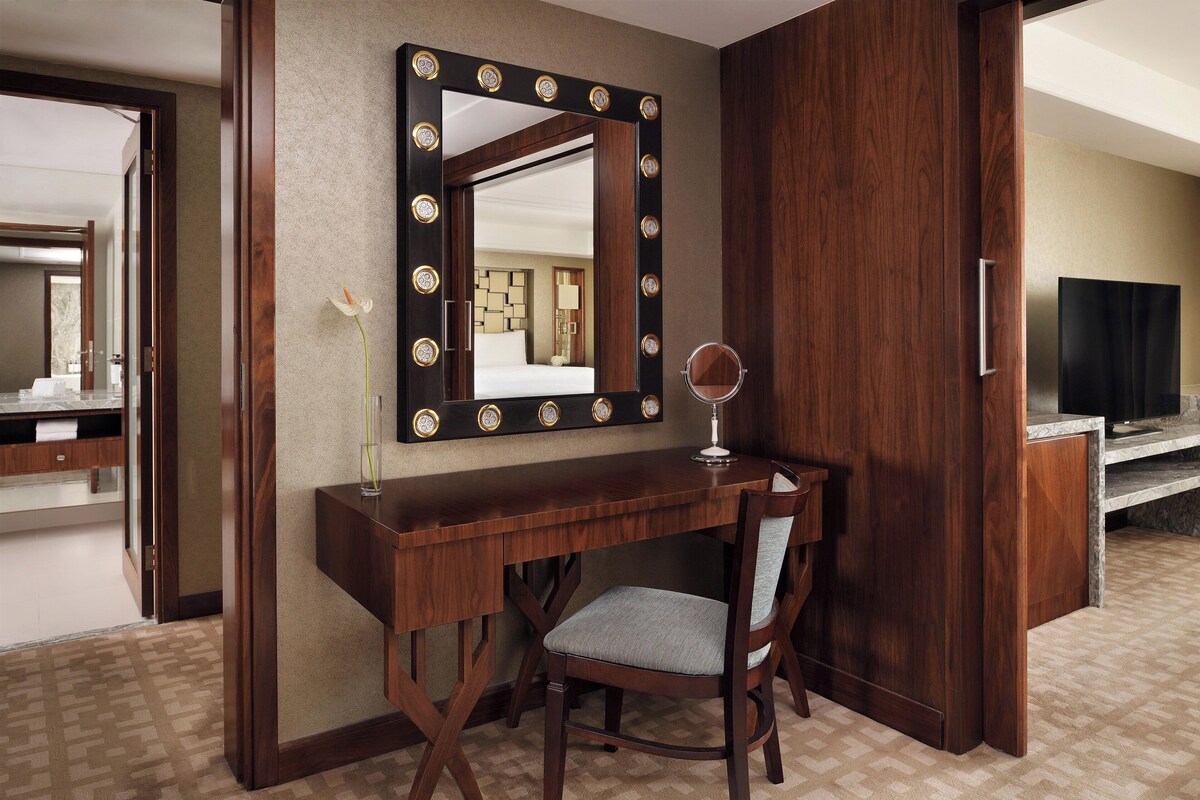 A hotel room with a vanity area featuring a mirror with lights, a wooden desk, and a chair.