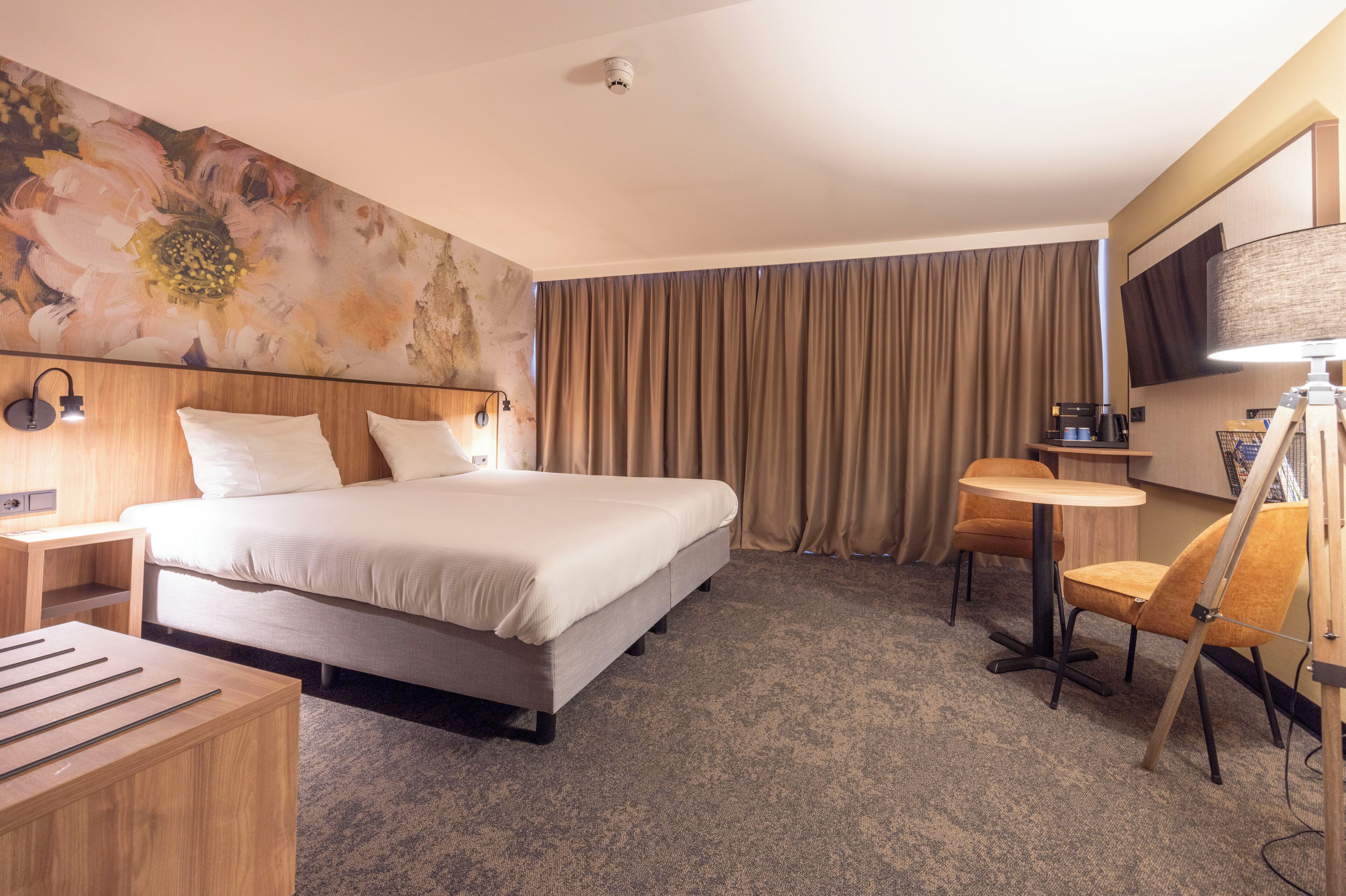 Quarto de hotel com uma cama, uma escrivaninha, uma cadeira, uma mesinha, uma televisão e um grande quadro floral na parede.