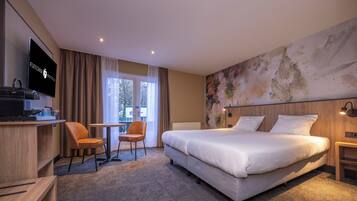 A hotel room with a large bed, a desk with a television, a chair, and a view of the outdoors through a window.
