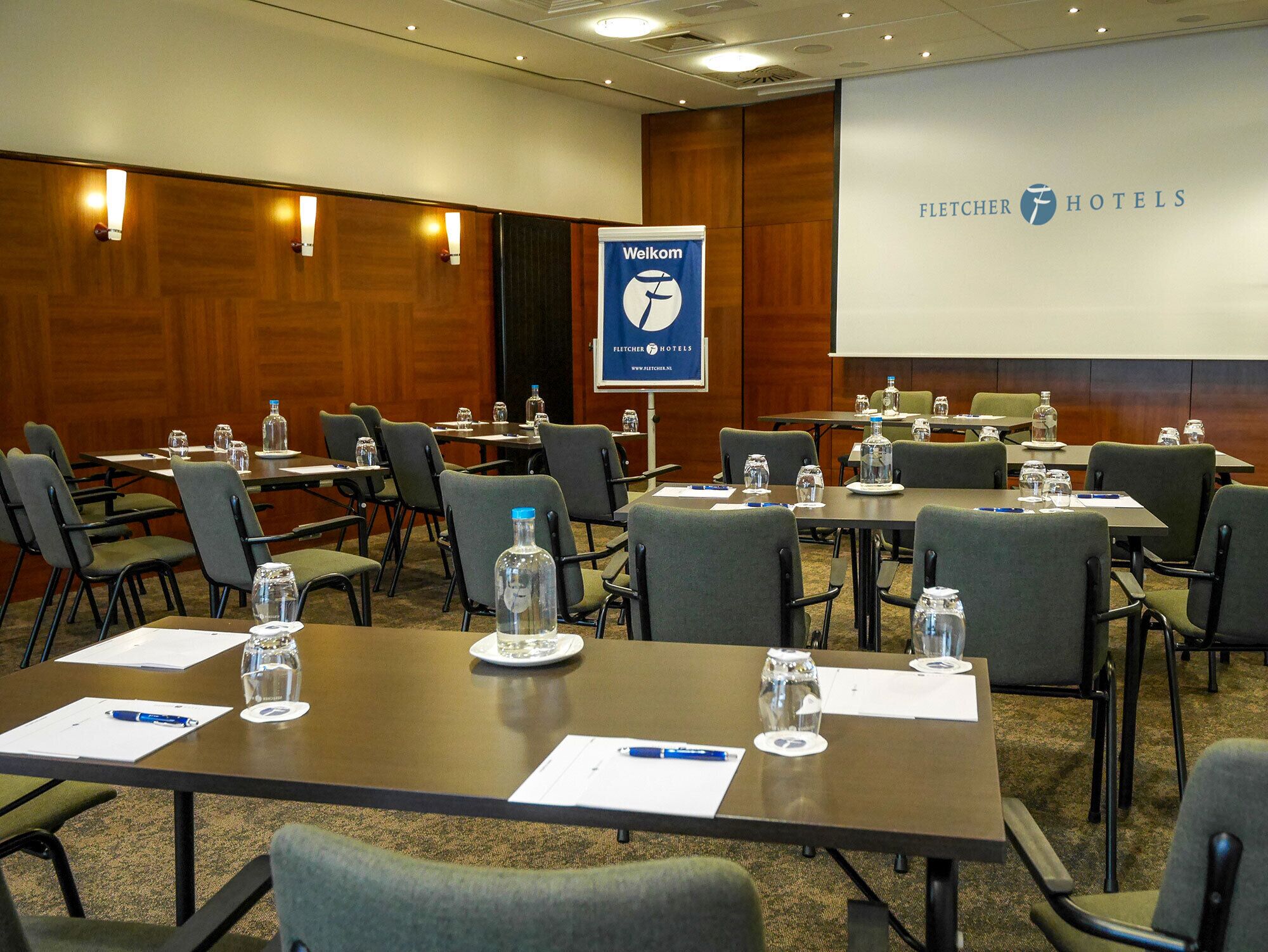 Una sala conferenze con tavoli, sedie, bottiglie d'acqua e un banner di benvenuto presso Fletcher Hotels.