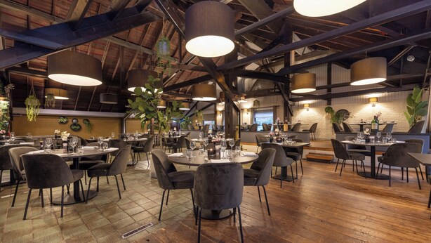 Un restaurante espacioso con techos altos, vigas a la vista y luminarias modernas. Las mesas están dispuestas con sillas, y el suelo es de madera.