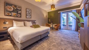 Una habitación de hotel con una cama grande, una mesita con sillas, un ventanal amplio y una planta.