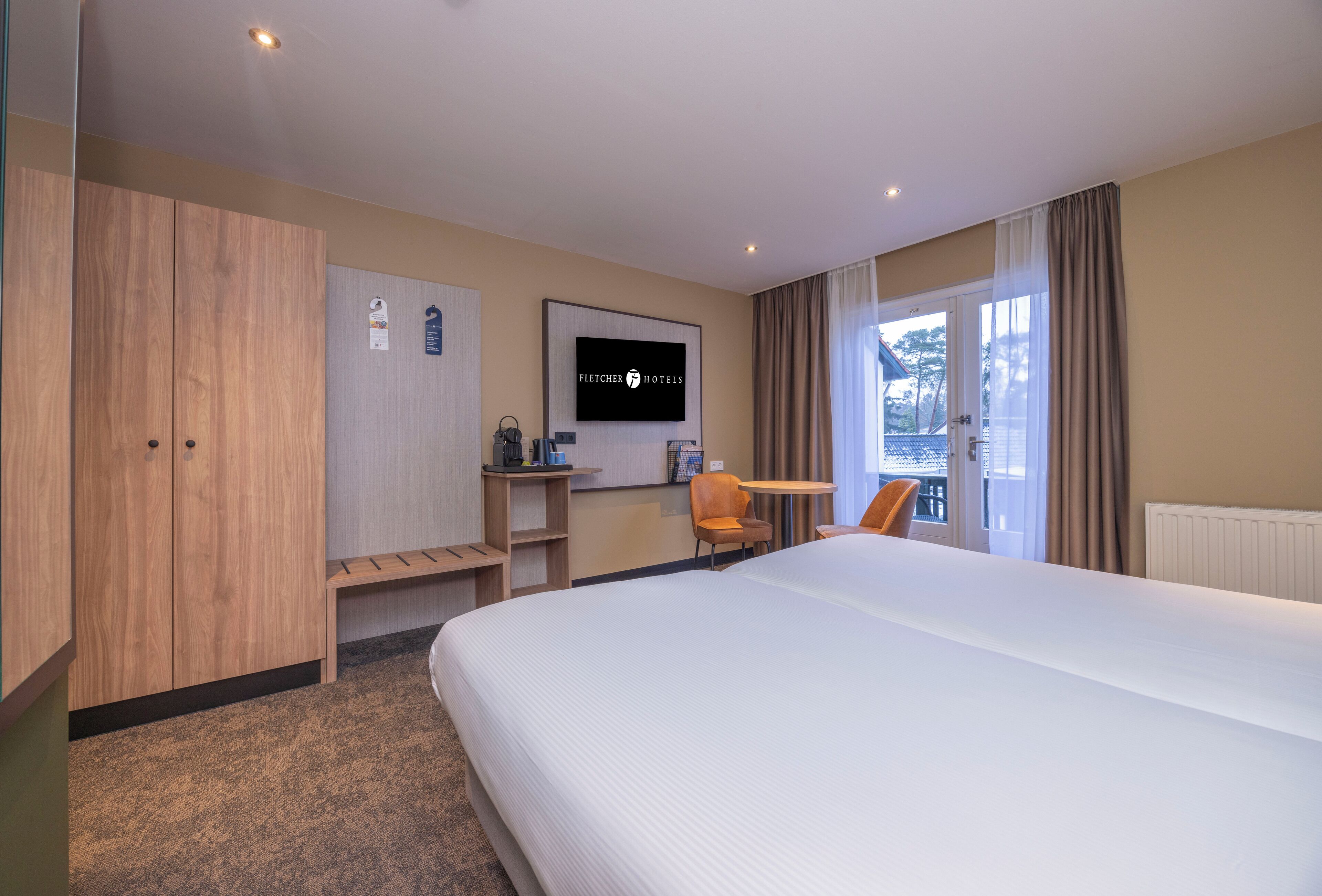 A hotel room with a large bed, wooden wardrobe, a desk with a chair, a television, and a window with curtains.